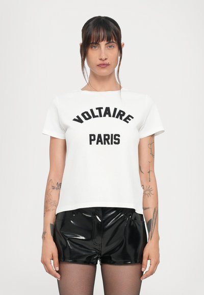 T-shirt in cotone bianco con "VOLTAIRE" e "PARIGI" stampati in grassetto nero. Maniche corte, scollatura rotonda. Abbinata a pantaloni corti neri lucidi.