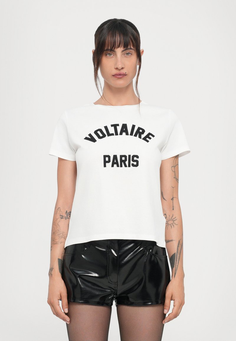Weißes Baumwoll-T-Shirt mit den Aufdrucken "VOLTAIRE" und "PARIS" in fettem Schwarz. Kurze Ärmel, runder Ausschnitt. Kombiniert mit glänzenden schwarzen Shorts.