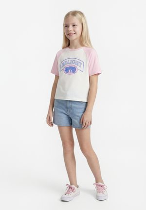 Egy fiatal lány áll és mosolyog, fehér és rózsaszín "DELIGHT" feliratú pólót, farmer rövidnadrágot és fehér, rózsaszín cipőfűzős tornacipőt visel.