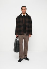 Jachetă din lână cu carouri maro, butoane negre, suprapusă peste un pulover din tricot negru. Pantaloni cu carouri maro, pantofi negri și o geantă tote neagră.