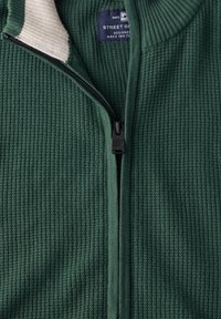 Maglione verde a coste con colletto color crema, dotato di chiusura con zip e etichetta con logo. Il tessuto ha un aspetto strutturato.
