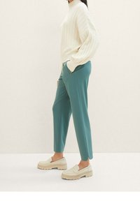 Gebreide crèmekleurige trui met hoge hals, gecombineerd met teal tapered broek en beige instap schoenen met een getextureerde zool, zijaanzicht.