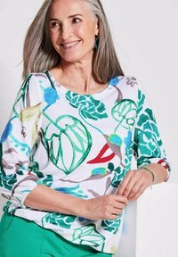 Witte longsleeve top met groene en blauwe botanische prints. Ronde halslijn en geraffineerde mouwen. Draagt met groene broek.