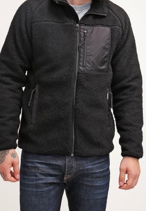 Man wearing a black fleece jacket with zippered chest and side pockets, paired with dark blue jeans against a plain background.