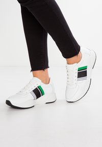 Białe sneakersy z teksturowaną cholewką, z czarnymi i zielonymi paskami oraz grubą podeszwą. Sznurowadła i język w kolorze białym. Czarna podeszwa zewnętrzna.