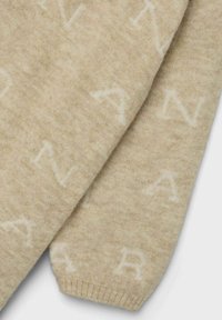 Neulepusero pehmeästä beige villasta, jossa on teksturoitu pinta ja toistuvia valkoisia kirjainkuvioita. Helman ribbaus takaa mukavan istuvuuden.
