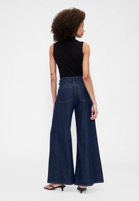 Temno modre kavbojke z visokim pasom in širokimi hlačnicami, s čistim zaključkom, ki imajo dva zadnja žepa in subtilne detajle šivov, kombinirane brezrokavnikom v črni barvi.