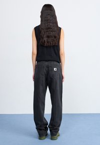 Top nero senza maniche e jeans larghi a righe grigio scuro. I jeans presentano tasche posteriori e un'etichetta di marca. Il modello indossa sneaker verdi.
