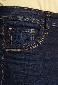 Donkerblauwe denim jeans met een zichtbare voorzak, contrasterende oranje stiksels en een gladde textuur. De tailleband is iets hoger.