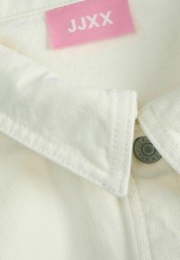 Camicia bianca con colletto realizzata in tessuto testurizzato, dotata di un'etichetta rosa con "JJXX" e un bottone metallico con design inciso.