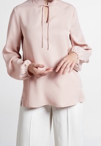 Helles rosa Oberteil mit hohem Kragen, Schlitzdetail, lockeren Ärmeln mit elastischen Bündchen und einer glatten, seidigen Textur. Seitenschlitze am Saum.