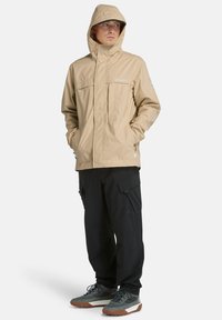 Beige vattentät jacka med huva, med två bröstfickor och elastiska ärmslut, bärs över svarta cargobyxor och grå skor.