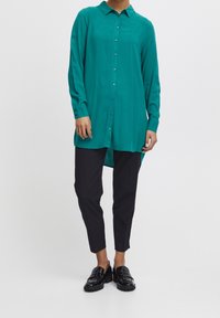 Chemise à manches longues bleu sarcelle avec col et boutons, en tissu lisse. Associée à un pantalon noir fuselé et des mocassins noirs brillants.
