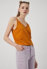 Oransje ermene topper med bølgete hals, sammen med lyse lilla jeans. Stoffet ser mykt ut med en lett teksturert finish.