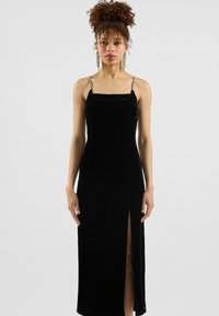 Žena u crnoj aksamitnoj haljini bez rukava s naramenicama od rhinestones i prorezom do butina, stoji nasuprot obične bijele pozadine.