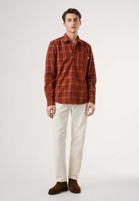 Camicia di flanella a quadri rossi e marroni con colletto a bottoni, taschino frontale e maniche lunghe, abbinata a pantaloni color crema e stivali marroni.
