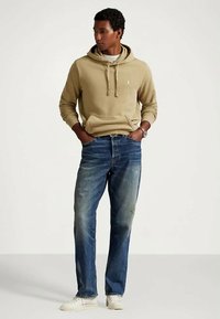 Άνδρας που στέκεται με τα χέρια στις τσέπες, φορώντας μπεζ φούτερ, μπλε τζιν και άσπρα αθλητικά παπούτσια, μπροστά από απλό φόντο.