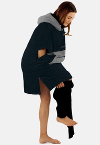 Dunkelblauer Kapuzenponcho mit grauen Akzenten und einer Fronttasche, ausgestattet mit seitlichen Schlitzen und einem strukturierten Stoff, der über einem Bein einer schwarzen Hose getragen wird.