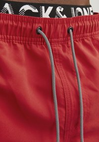 Rode zwemshorts met een trekkoordsluiting. Zwarte tailleband met witte tekst "JACK & JONES". Glad, lichtgewicht materiaal met een matte afwerking.