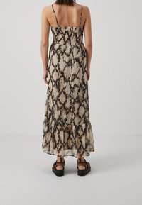 Nainen, joka on pukeutunut hiattomaan, nilkkapituiseen mekkoon musta-beige sitruunadipesu-kuvioinnilla, yhdistettynä ruskeisiin sandaaleihin, seisoo yksinkertaista taustaa vasten.