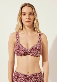 Burgunderfärgad mönstrad bikinitopp med vriden frontdesign, krämfärgade blommotiven och vågformig kant längs kanterna. Justerbara axelband.