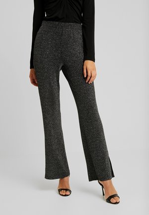 Frau trägt funkelnde dunkelgraue weit geschnittene Hosen mit Seitenschlitzen, schwarzes Langarmshirt und schwarze offene High Heels, steht auf hellem Hintergrund.