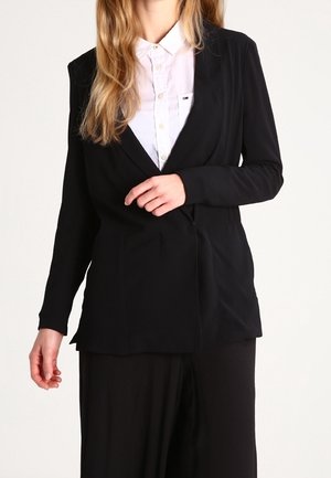 Frau trägt einen schwarzen Blazer über einem weißen Hemd, richtet den Blazer mit einer Hand, neutraler Hintergrund.