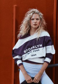 Biely a fialový oversize sveter s nápisom "BROOKLYN NEW YORK 1994", s rebrovaným golierom a manžetami; v kombinácii so svetlomodrými legínami.
