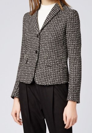 Woman wearing a black and white tweed blazer over a white top paired with black pants, against a plain light gray background.