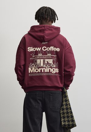Mand med flettet hår vendt væk iført en mørkerød hættetrøje med teksten "Slow Coffee Mornings" og en caféillustration på bagsiden.