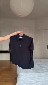 Ręka trzyma czarną koszulę z krótkim rękawem i kołnierzykiem na wieszaku w jasnej sypialni z białym łóżkiem i drewnianą podłogą.