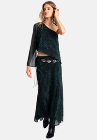 Vestido verde estampado con escote asimétrico, mangas largas transparentes y un cinturón decorativo plateado. Combinado con botas negras hasta el tobillo.