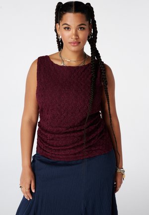 Femme aux longs cheveux tressés portant un haut bordeaux texturé sans manches, une jupe bleu marine, des créoles et des colliers superposés, debout devant un fond blanc.