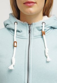 Felpa con cappuccio azzurro chiaro, con un interno morbido e testurizzato, dotata di zip in metallo, lacci in cotone e dettagli in legno nei punti di gomma.