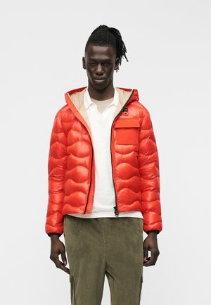 Bărbat cu piele închisă la culoare și dreadlocks, purtând o geacă puf portocalie deschisă peste un tricou alb și pantaloni verde măsliniu, stând în picioare pe fundal alb.