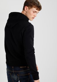 Felpa nera con tasca a marsupio e cappuccio con cordino, indossata con jeans blu con cuciture arancioni. Tessuto misto cotone, vestibilità casual.