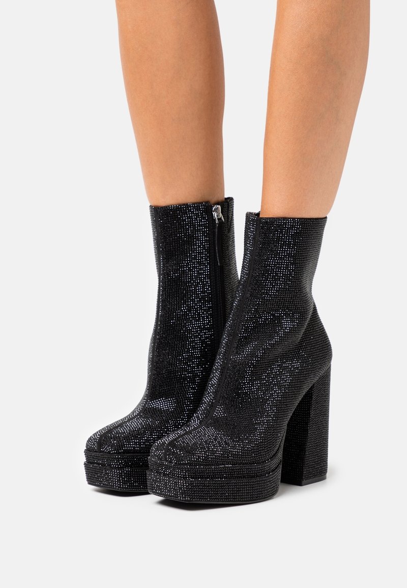 Jambes inférieures portant des bottines noires à plateforme recouvertes de petits strass scintillants avec fermeture éclair latérale et talons carrés.