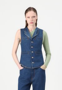 Denim brezrokavnik s prilagojeno zasnovo, zarezan ovratnik in zapiranje na pet gumbov. Ima dve žepek z apnjenjem v oranžni barvi. Nosite ga čez zeleno majico.