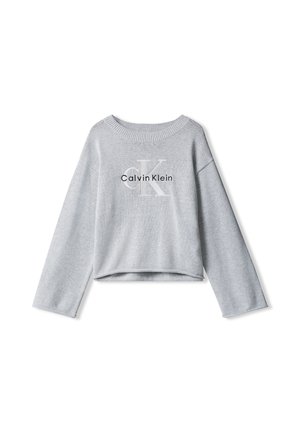 Dziecko z długimi włosami nosi szarą bluzę Calvin Klein, czarne spodnie, czarny sweter na ramionach oraz czarne masywne buty, stoi nieruchomo.