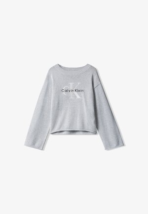 Enfant aux longs cheveux porte un sweat-shirt gris Calvin Klein, un pantalon noir, un pull noir posé sur les épaules et des chaussures noires épaisses, debout immobile.