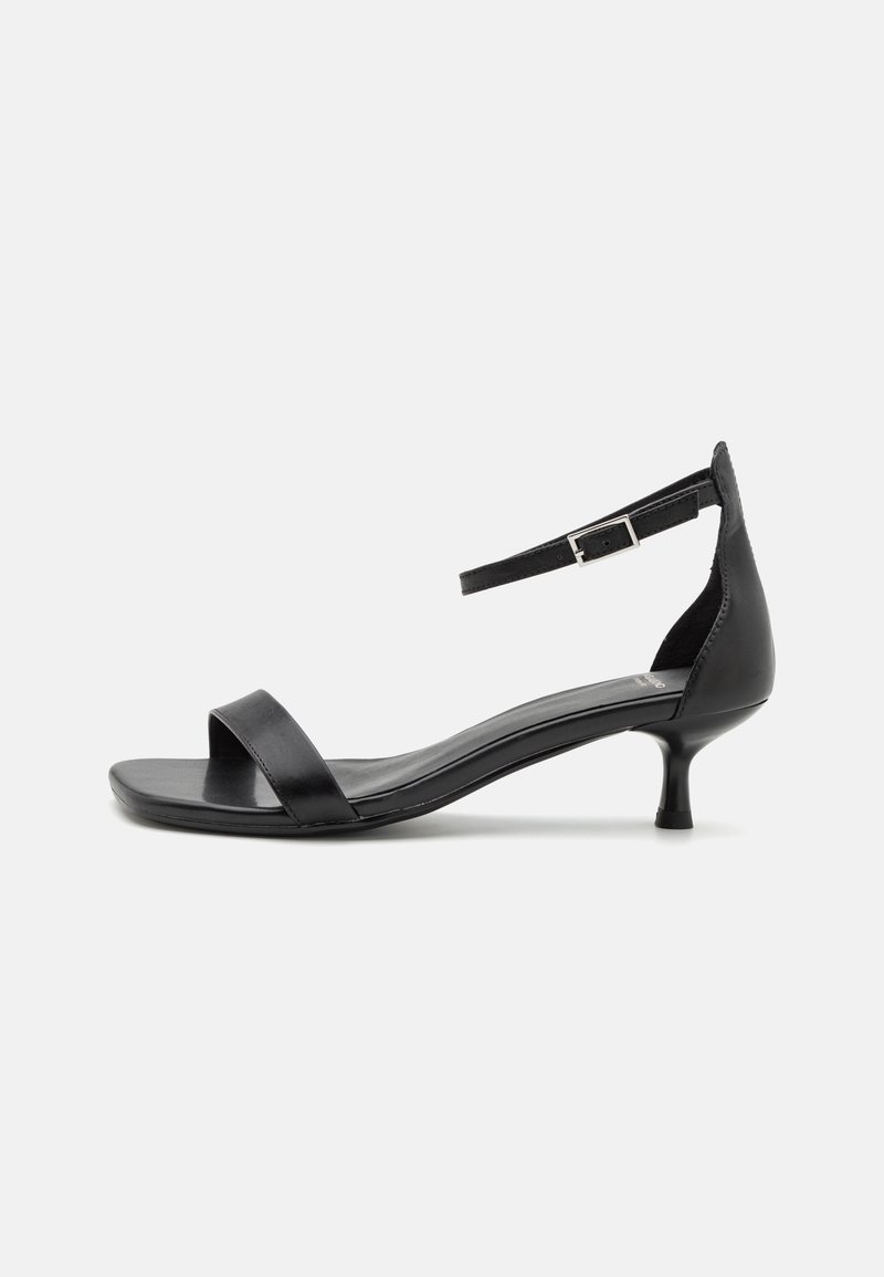 Sandale noire à petit talon avec une fine bride avant et une bride ajustable à la cheville avec une boucle argentée sur fond blanc.