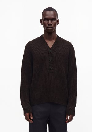Man draagt een donkerbruine geribde gebreide trui met drie knopen bij de V-hals en een donkere broek tegen een effen witte achtergrond.