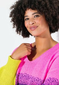 Maglione overdimensionato di un brillante colore rosa con scollatura a V, caratterizzato da polsini a costine gialli e una sezione centrale decorata con tonalità di viola. Texture morbida.