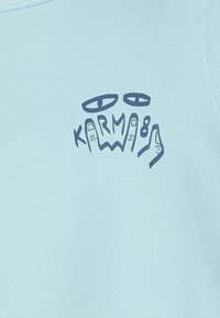 Gaiši zils krekls ar tumši zilu grafisko dizainu, kas attēlo rokas, acis un uzdrukātu tekstu "KARM8" uz priekšpuses.