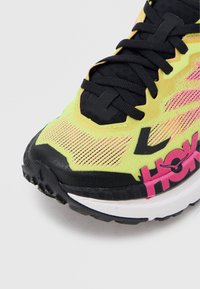 Primo piano della scarpa da corsa gialla e rosa con lacci neri, puntale nero e parte del logo rosso "HOKA" sulla suola bianca.