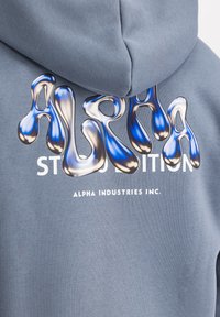 Felpa blu con lettering in rilievo effetto scioglimento "ALPHA" in argento e blu. Sotto, testo bianco che recita "STUDIO EDITION ALPHA INDUSTRIES INC."