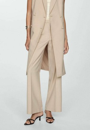 Pantaloni beige su misura abbinati a un lungo gilet beige, con bottoni doppi e uno spacco laterale, completati da sandali con tacco neri.