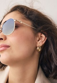 Un paio di occhiali da sole tono oro con lenti azzurro chiaro, abbinati a un orecchino dorato lucido. L'orecchino ha un design liscio e arrotondato.