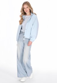 Vaaleansininen bomber-takki raidallisen paidan päällä, yhdistettynä leveälahkeisiin vaaleanpesuisiin farkkuihin ja valkoisiin tennareihin. Sileä kangas ja rento istuvuus.