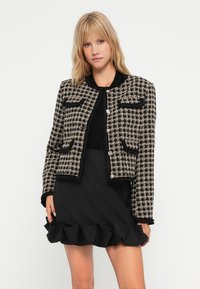 Tweedjakke i svart og kremmønstret rutete design, med to frontlommer, rund halslinje og svart kant. Modellen har på seg et volangskjørt i svart.